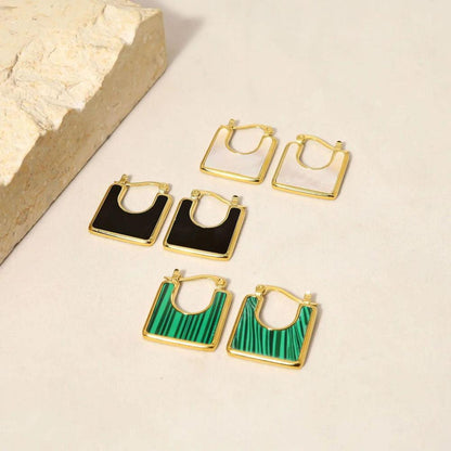 A set of square-shaped gold-plated earrings with enamel detailing in black, white, and green, displayed on a neutral background with a textured stone accent.