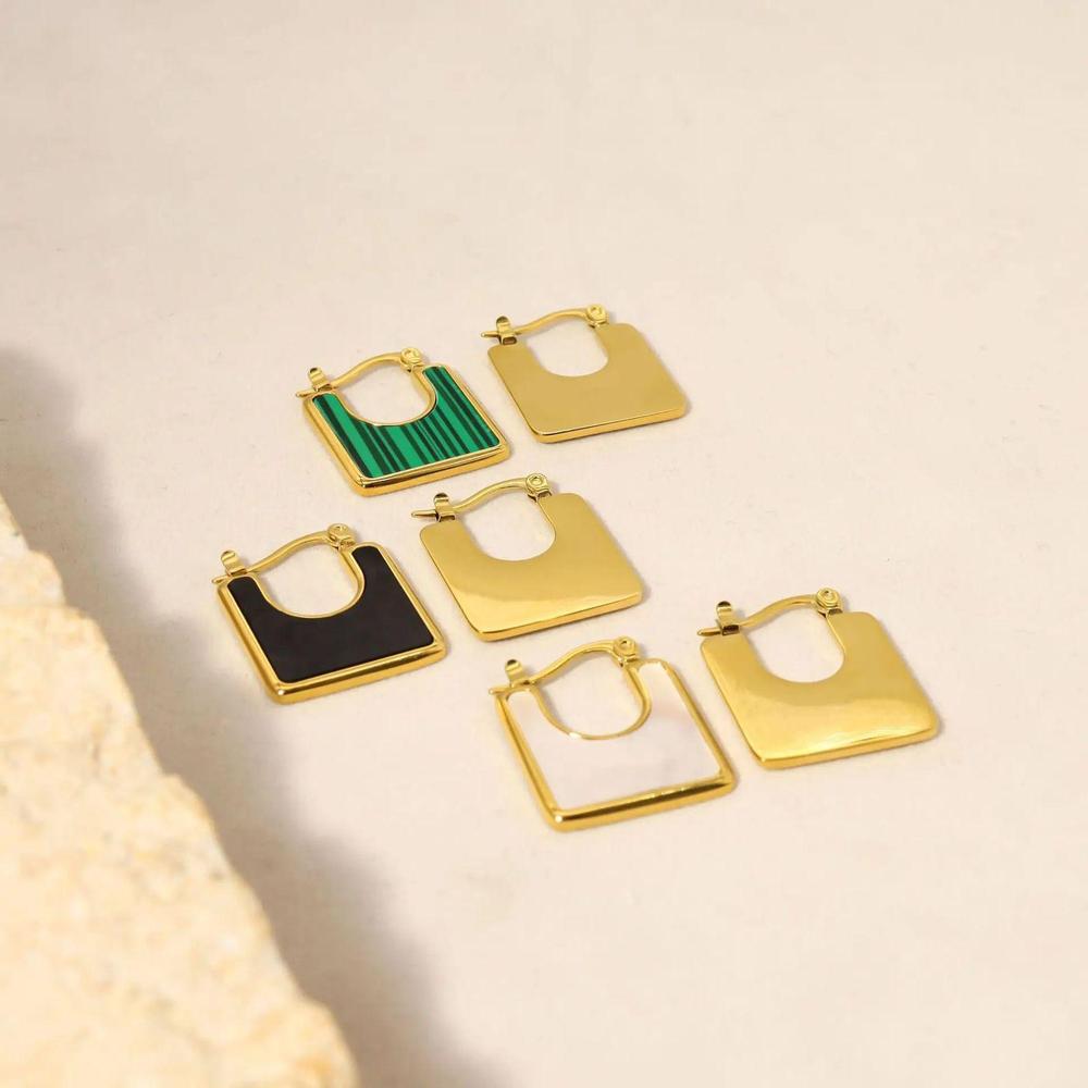 A set of square-shaped gold-plated earrings with enamel detailing in black, white, and green, displayed on a neutral background with a textured stone accent.